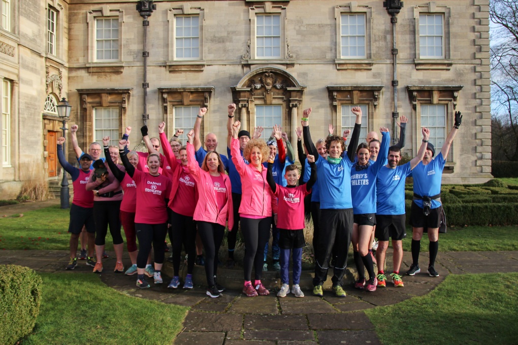 A group of people wearing pink Fitmums & Friends or blue Curly's Athletes tops in front of Normanby Hall. They are raising their arms in a cheer.