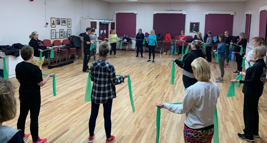 A group of women standing in a circle in a room. They are using resistance bands to do an upper-body exercise.